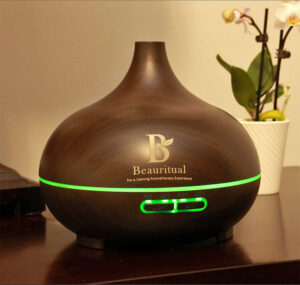 Dark wood ultrasonic essential oil diffuser by Beauritual emitting calming green light with orchid in background.
