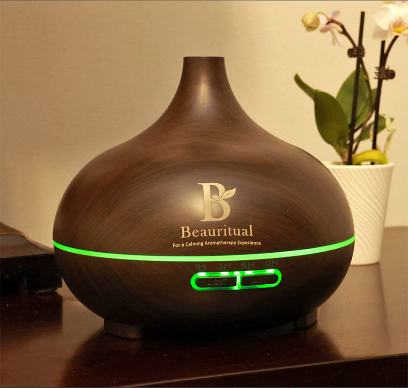 Dark wood ultrasonic essential oil diffuser by Beauritual emitting calming green light with orchid in background.